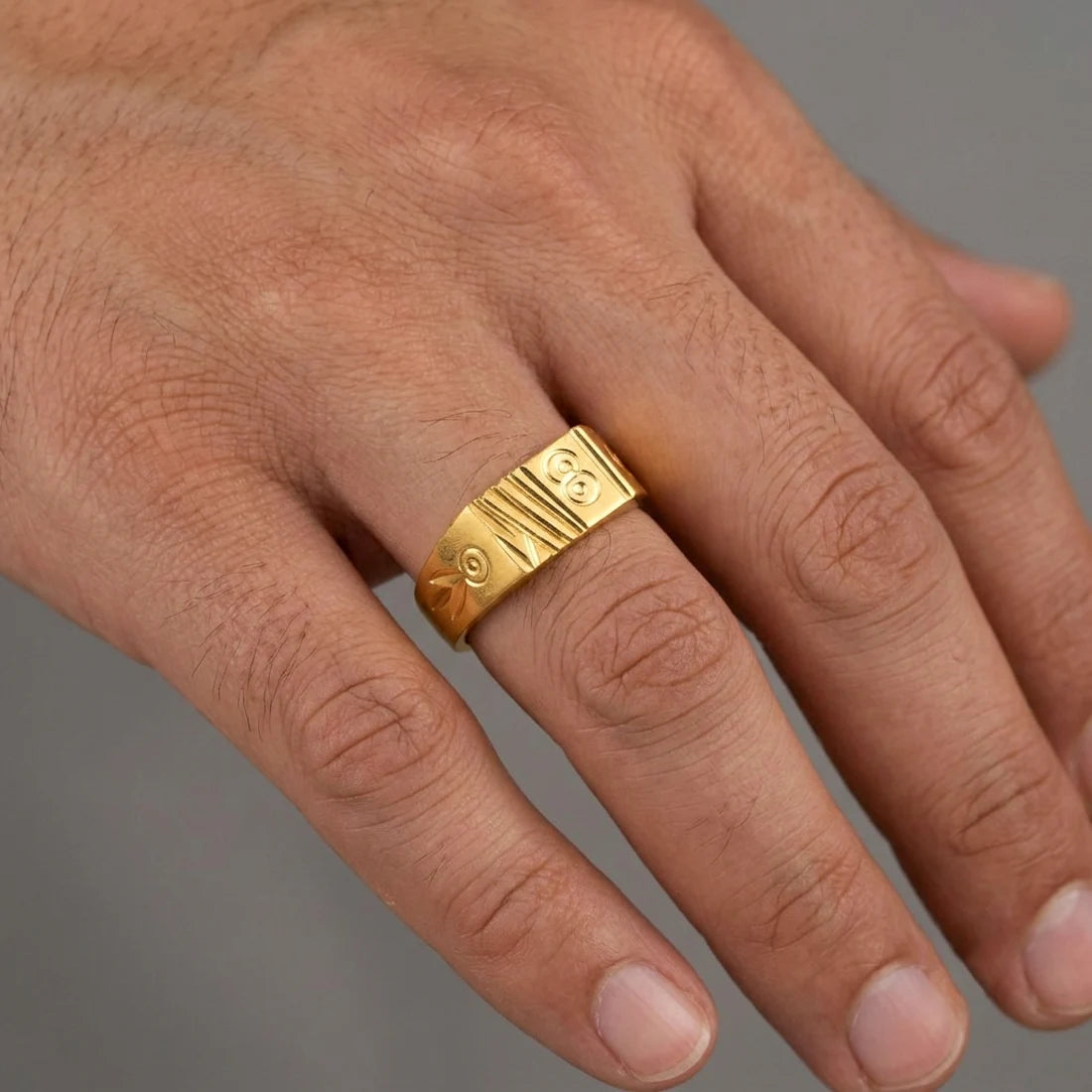 A close-up of a hand wearing a wide, polished yellow gold ring with engraved geometric and circular patterns on the band, set against a neutral gray background.