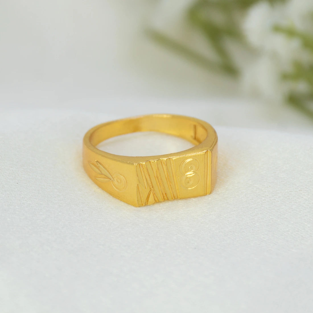 A gold signet-style ring with engraved decorative details, resting on a soft white textured surface with a blurred white flower in the background.