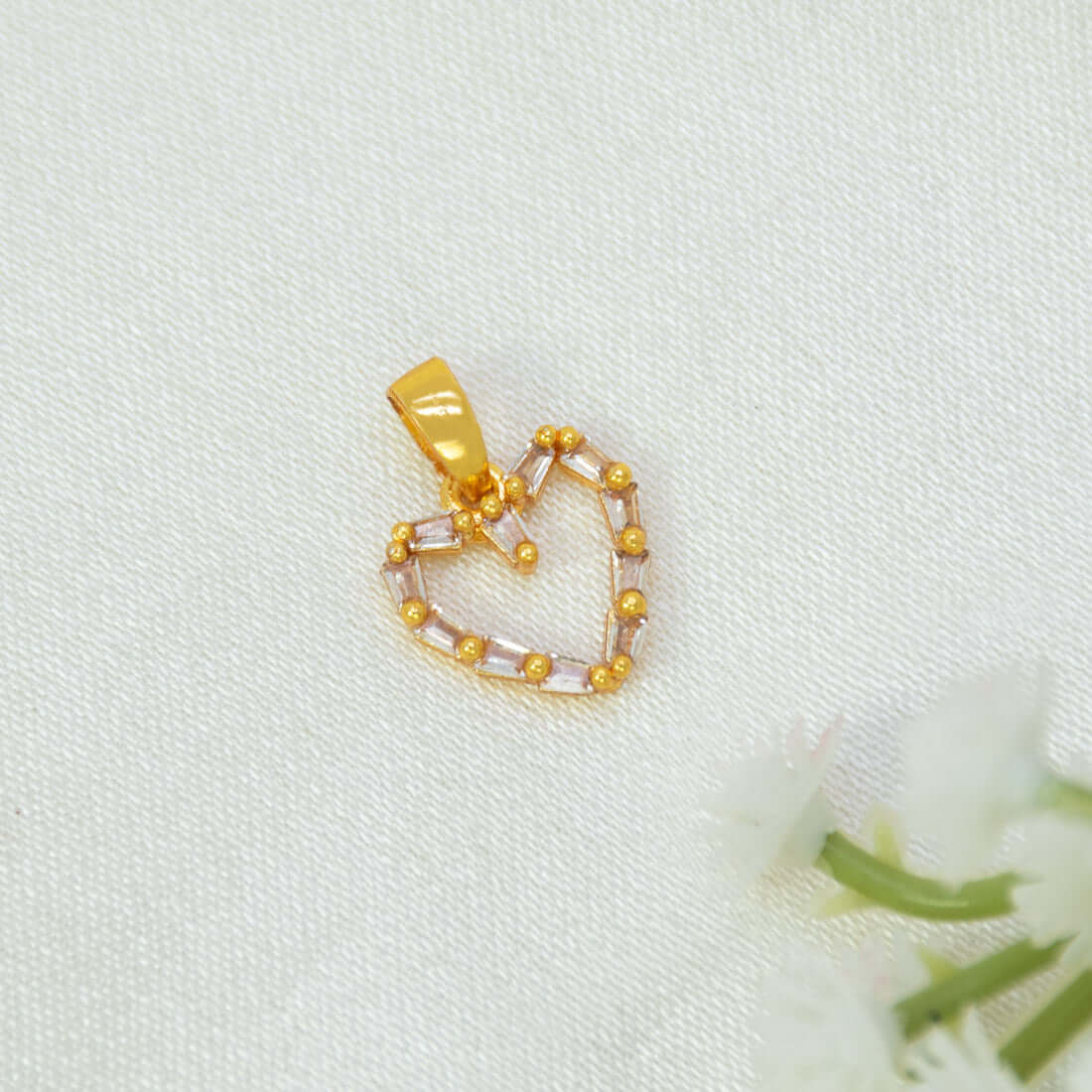 A Gold-Plated Baguette-Cut Cubic Zirconia Heart Pendant with clear rectangular CZ stone and small gold beads, resting on a textured white fabric background with white flowers in the corner.