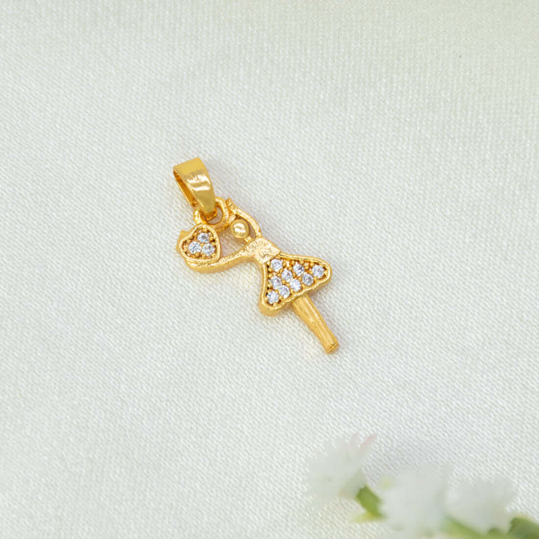 A gold-plated ballerina dancer pendant adorned with clear rhinestones, resting on a textured white fabric background with small white flowers in the corner.