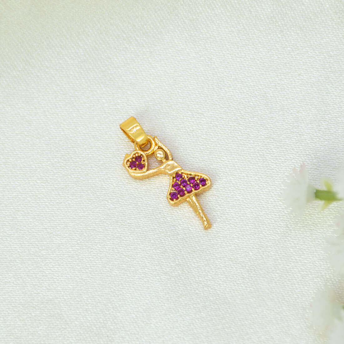 A gold-plated ballerina dancer pendant  with a ruby stone heart and purple diamond-patterned dress, resting on a textured white fabric background.