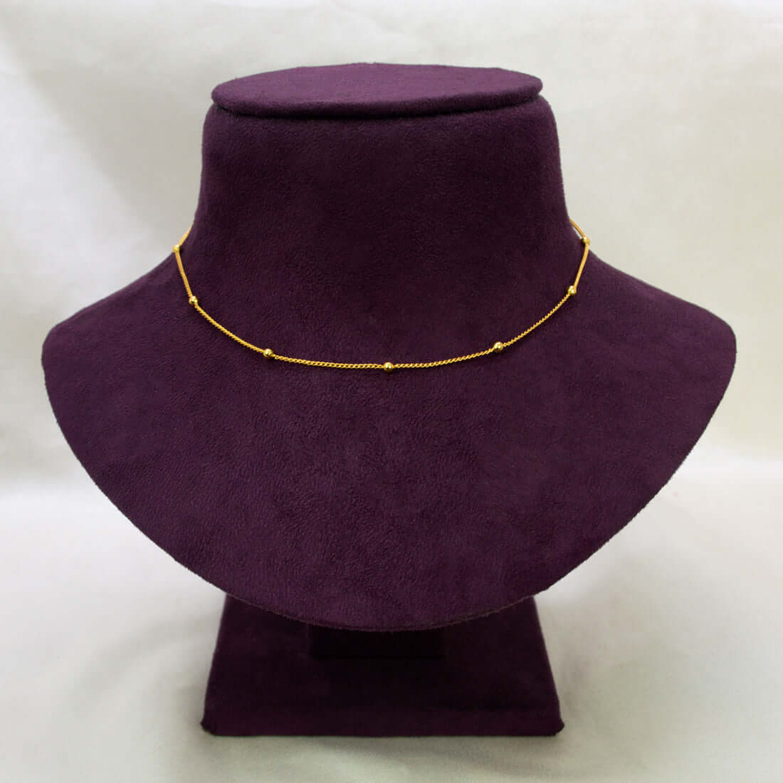 A Gold-Plated Beaded Curb Link Baby Chain necklace with small bead-like links, displayed on a dark purple velvet jewelry bust against a light off-white background.