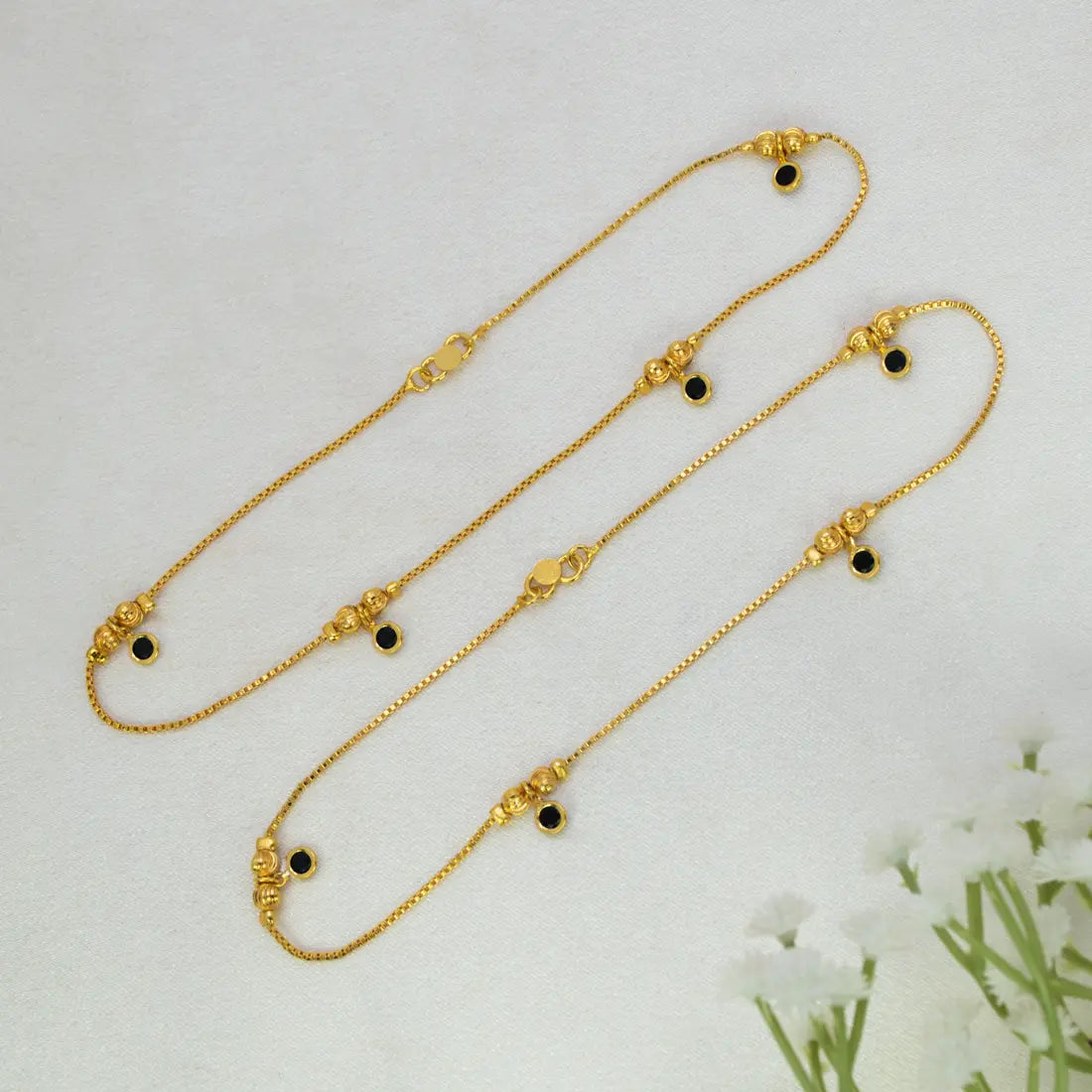 Gold-Plated Black Crystal Gold Bead Anklets with small black circular stones and decorative gold beads, arranged on a light textured background with white flowers in the corner.