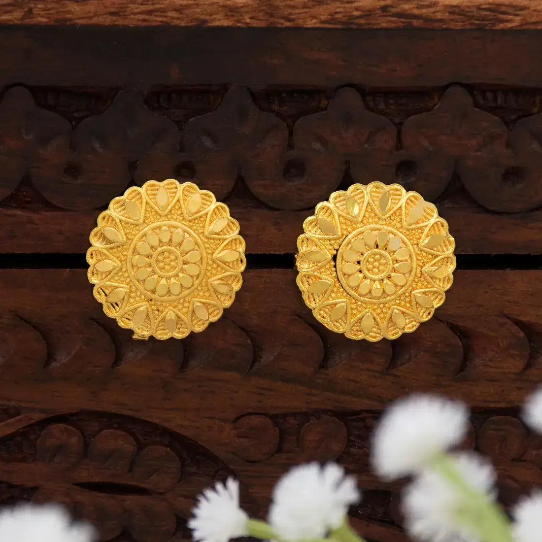 A pair of Gold-Plated Blooming Heart Stud Earrings For women with intricate filigree and floral mandala designs, displayed on a dark carved wooden surface with blurred white flowers in the foreground.