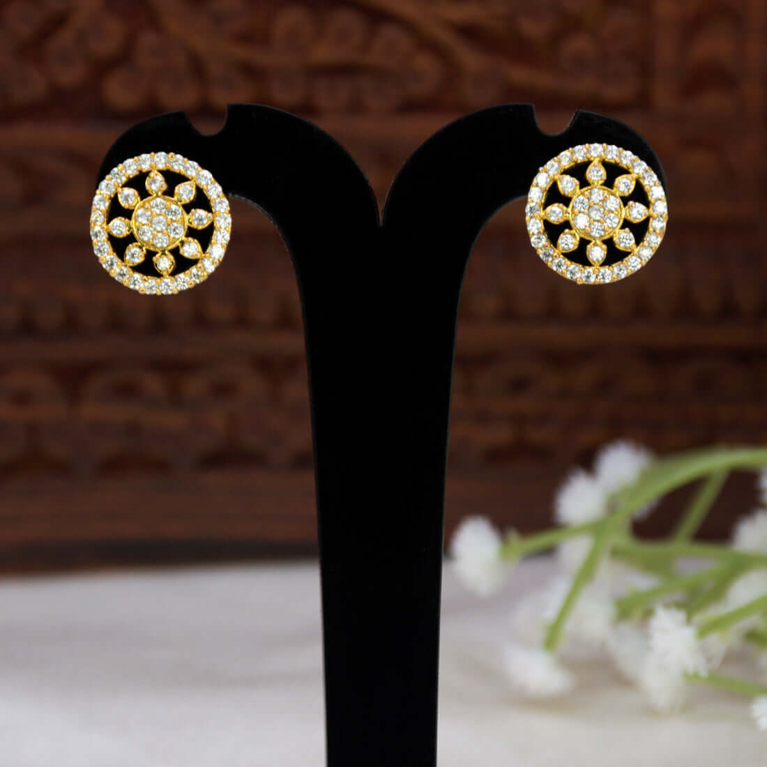 A pair of Gold-Plated CZ Sunburst Stud Earrings with clear pavé-set stones, displayed on a black T-shaped stand against a dark carved wooden background with blurred white flowers in the foreground.