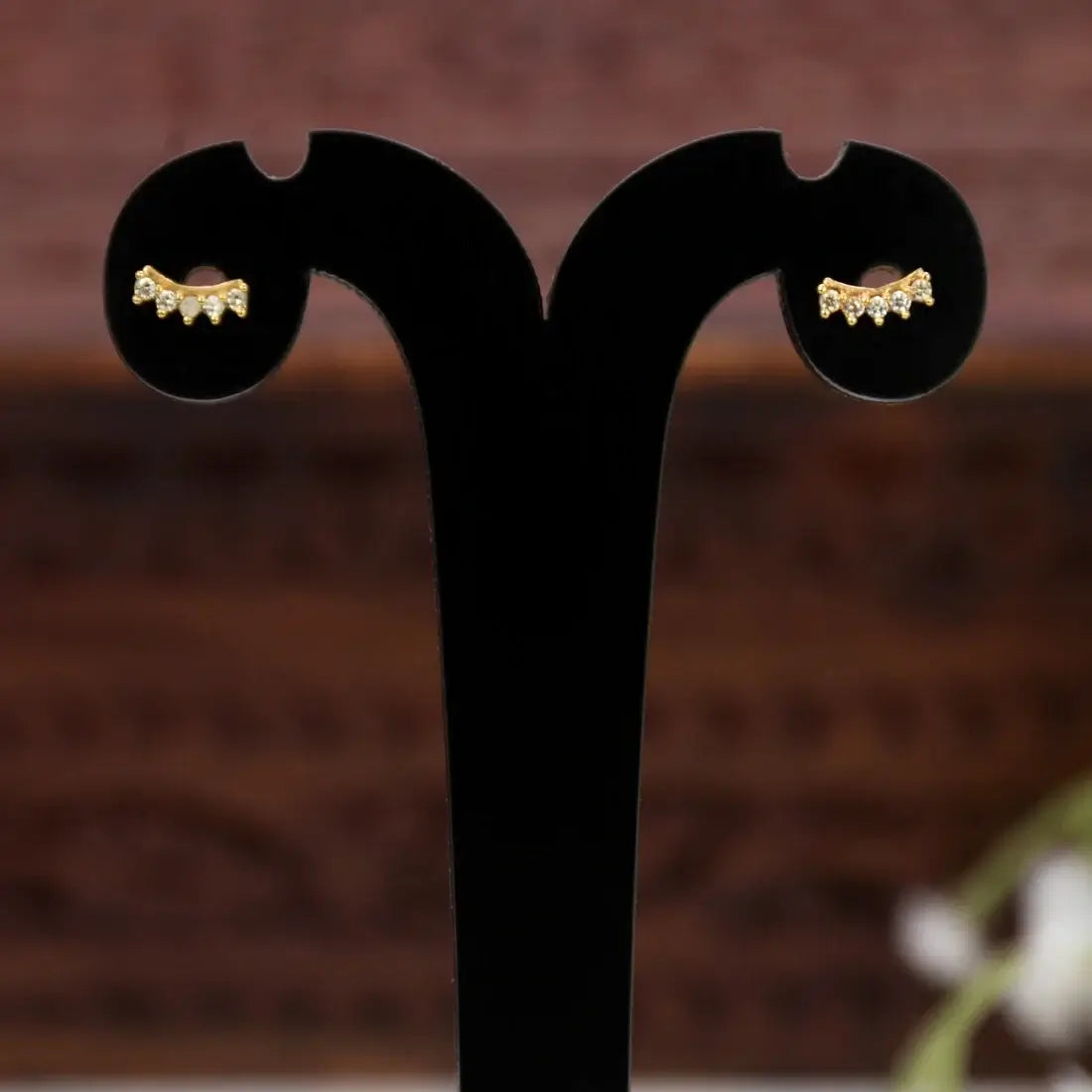 A pair of Gold-Plated Cubic Zirconia Arc Second Stud Earrings with tiny clear stones, displayed on a black T-shaped jewelry stand against a blurred brown background.