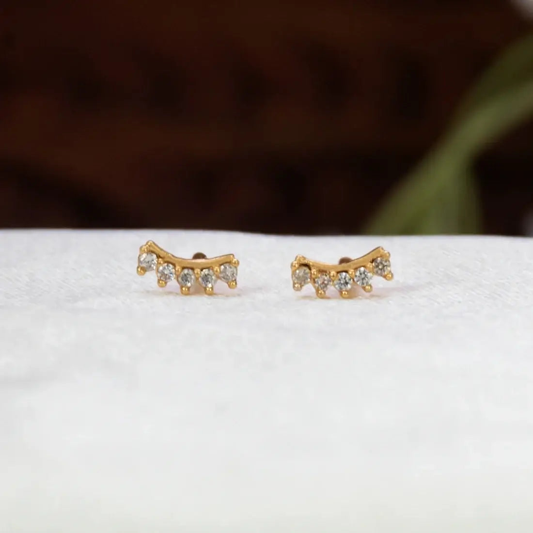 A pair of Gold-Plated Cubic Zirconia Arc Second Stud Earrings adorned with small clear gemstones, displayed on a textured white surface with a blurred dark brown background.