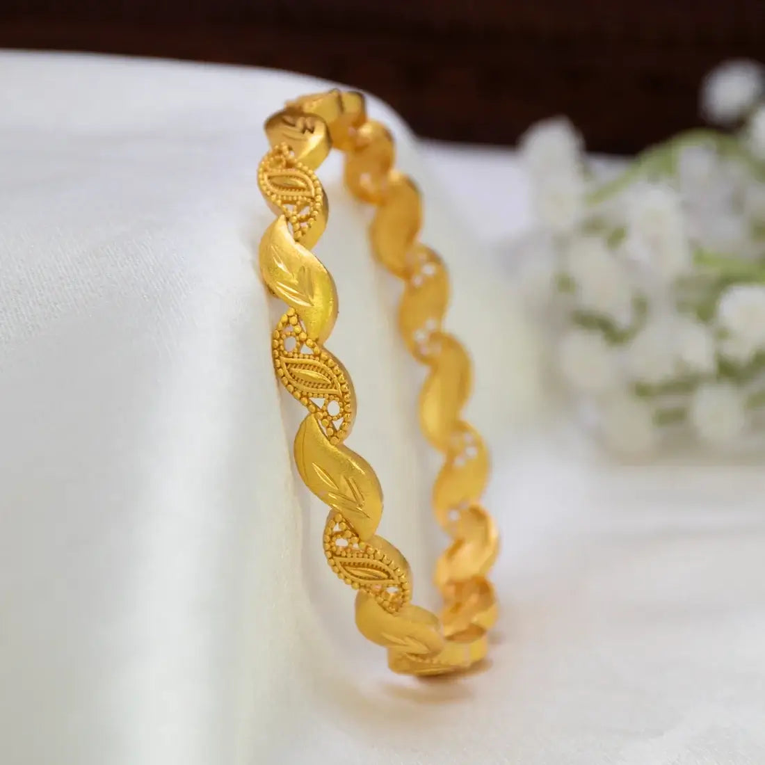 A gold-plated designer leaf bangle bracelet with a twisted leaf pattern displayed on a white fabric surface with a blurred floral background.