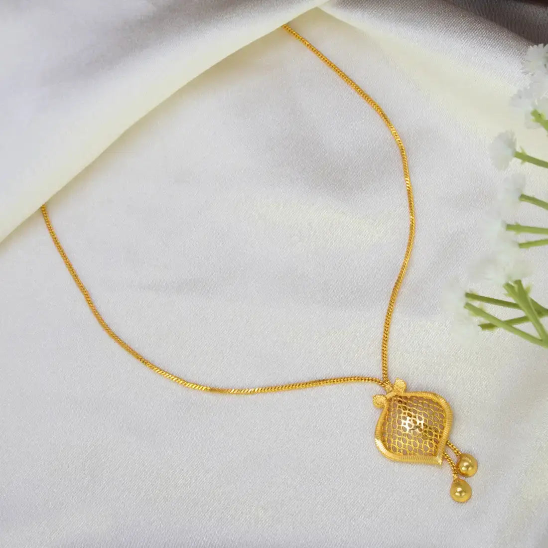 A Gold-Plated Filigree Mesh Pendant Chain with a decorative gold pendant, styled on a draped white fabric background with small white flowers on the side.