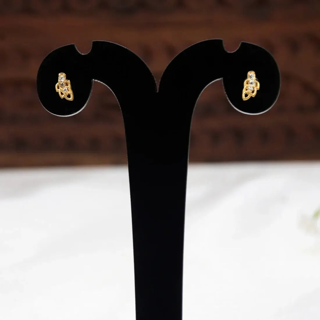 A pair of Gold-Plated Leaf Design CZ Stone Second Studs displayed on a black T-shaped jewellery stand against a blurred brown and white background.