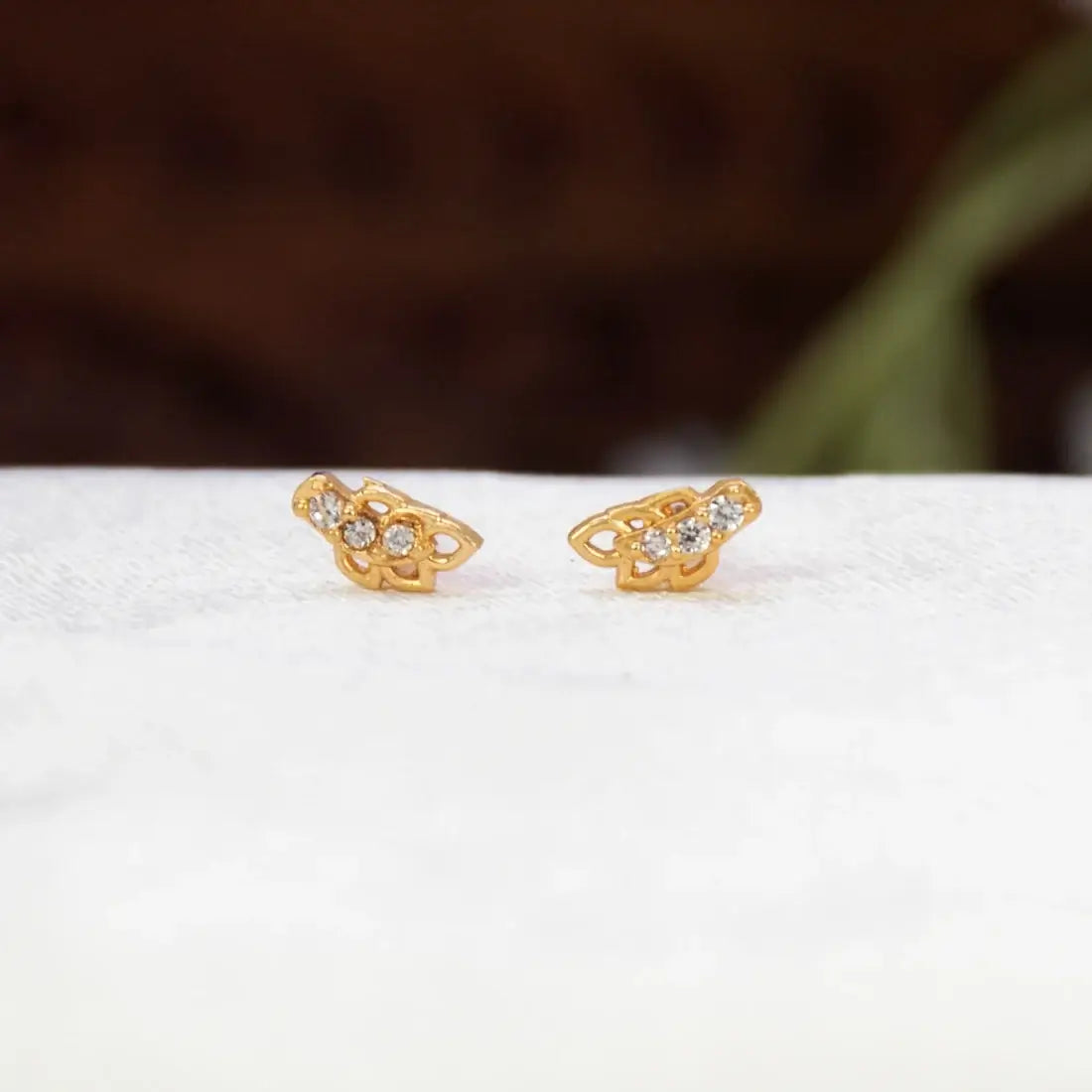 A pair of Gold-Plated Leaf Design CZ Stone Second Studs with small clear stones, displayed on a white textured surface with a blurred dark brown background.