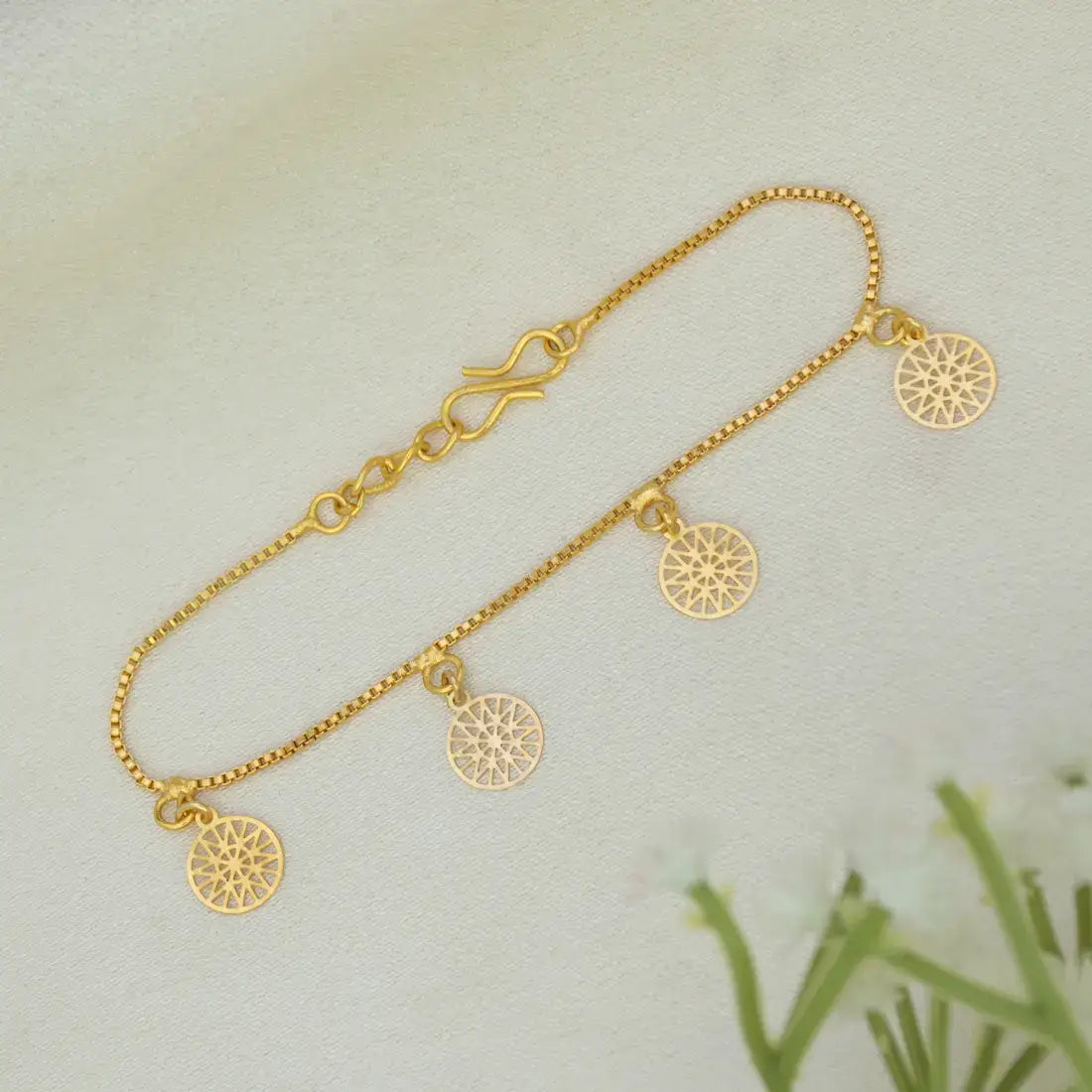 A delicate Gold-Plated Mandala Charm Hanging Chain Bracelet with four circular sunburst-style charms, displayed on a soft, light beige textured surface with a hint of green leaves in the corner.