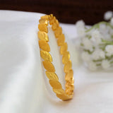 A Gold-Plated Oval Leaf Design Bangle displayed on a white fabric surface with small white flowers in the background.