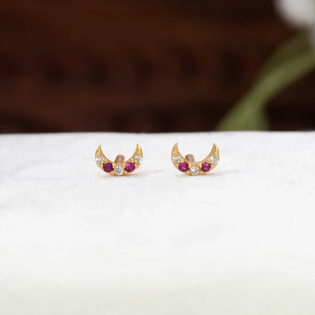 A pair of Gold-Plated Ruby CZ Crescent Second Stud Earrings with small red gemstones and clear stones, displayed on a white textured surface with a blurred dark brown background.