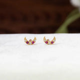 A pair of Gold-Plated Ruby CZ Crescent Second Stud Earrings with small red gemstones and clear stones, displayed on a white textured surface with a blurred dark brown background.