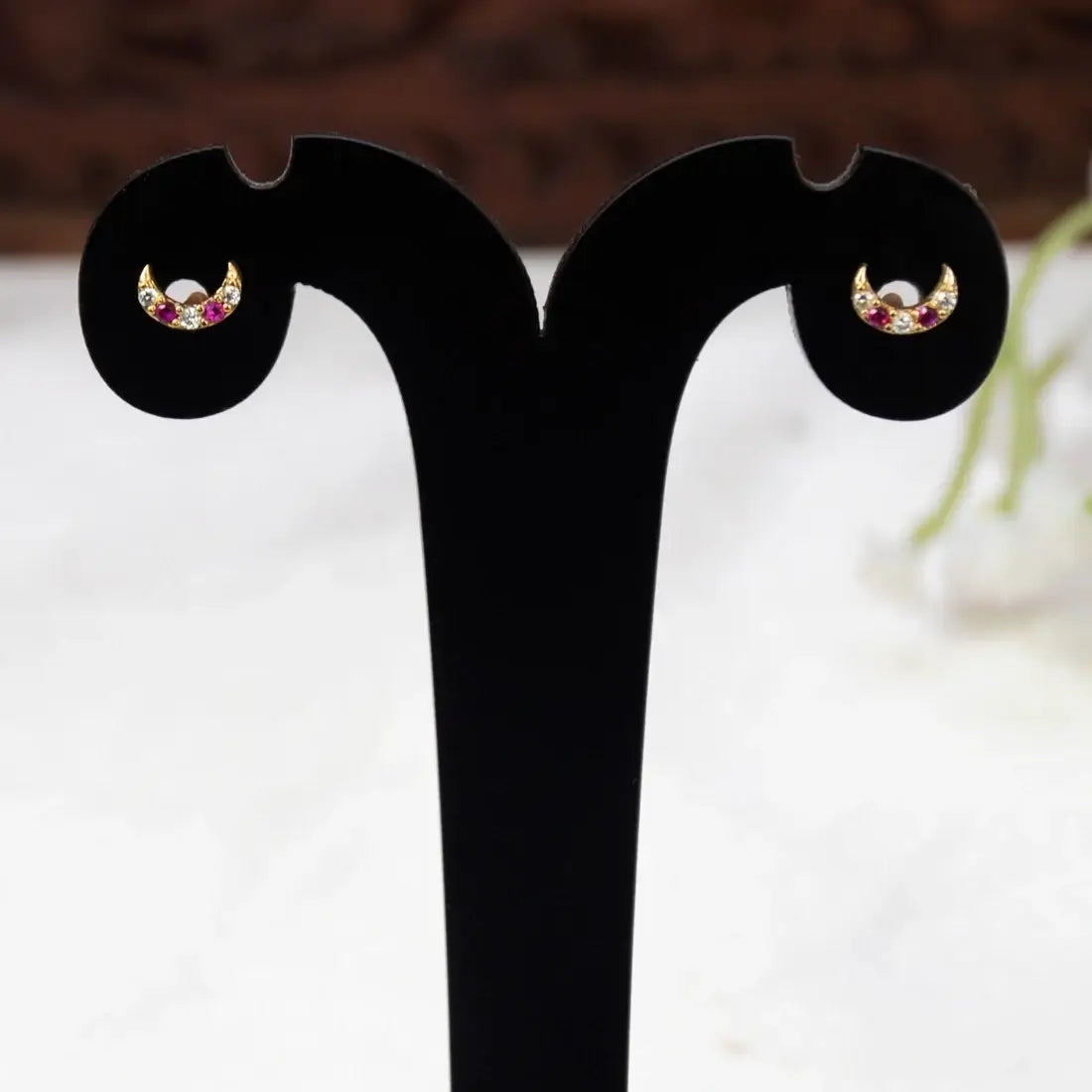 A pair of Gold-Plated Ruby CZ Crescent Second Stud Earrings with small red and white gemstones, displayed on a black T-shaped jewelry stand against a blurred white and brown background.