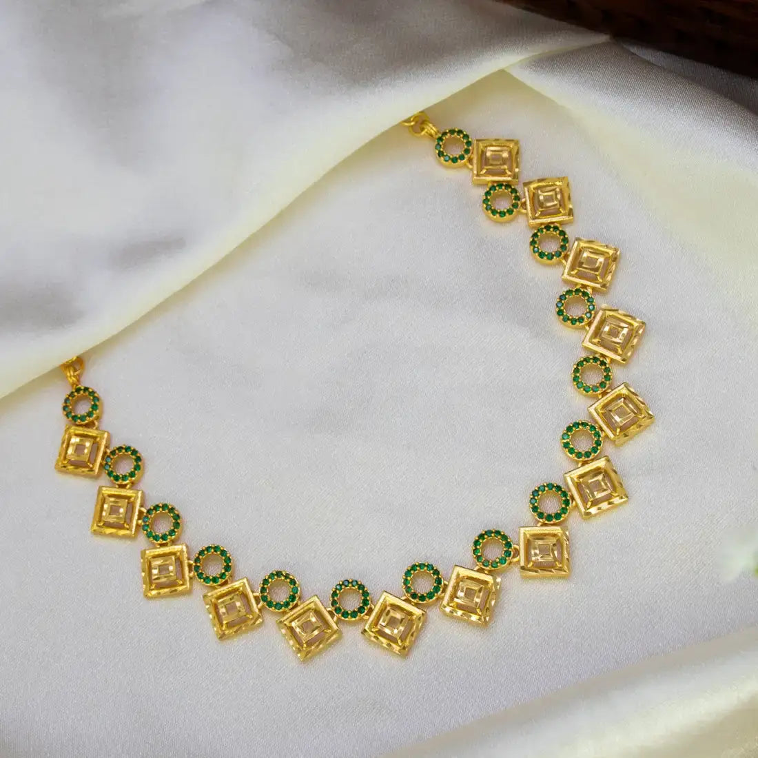 A gold-plated necklace featuring alternating square and circular links, with the circular links accented by small green stones, displayed on a draped white satin fabric.