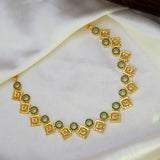 A gold-plated necklace featuring alternating square and circular links, with the circular links accented by small green stones, displayed on a draped white satin fabric.