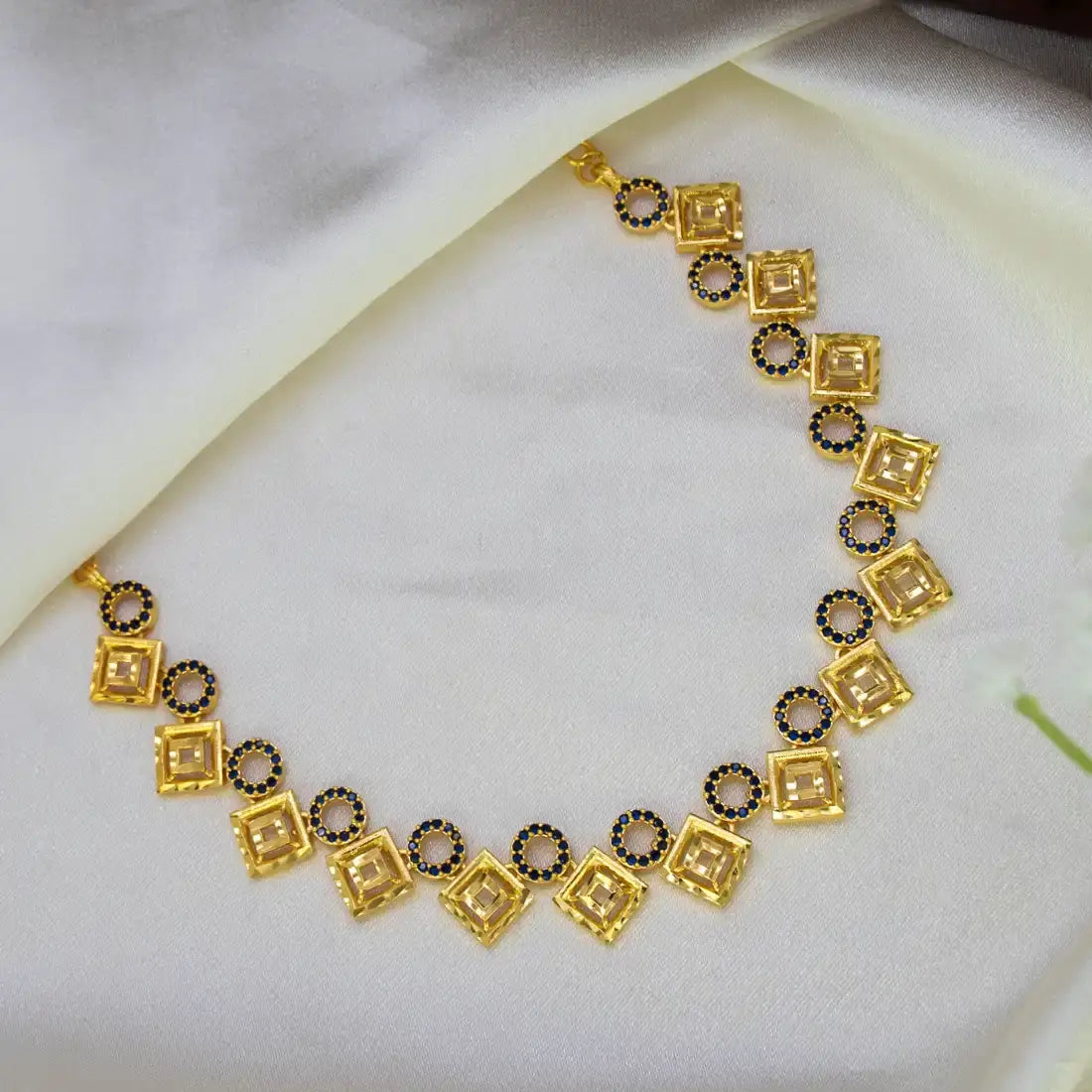 A Gold Plated Square-and-Circle Stone Necklace featuring alternating square and circular motifs, with clear rectangular stones in the centers and small dark blue accents, displayed on a draped white satin fabric.