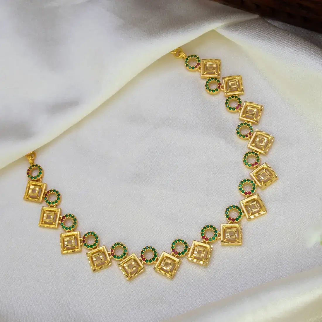 A Gold Plated Square-and-Circle Stone Necklace featuring square and circular motifs, embellished with small green and pink stones, displayed on a draped white satin fabric.