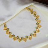 A Gold Plated Square-and-Circle Stone Necklace featuring square and circular motifs, embellished with small green and pink stones, displayed on a draped white satin fabric.