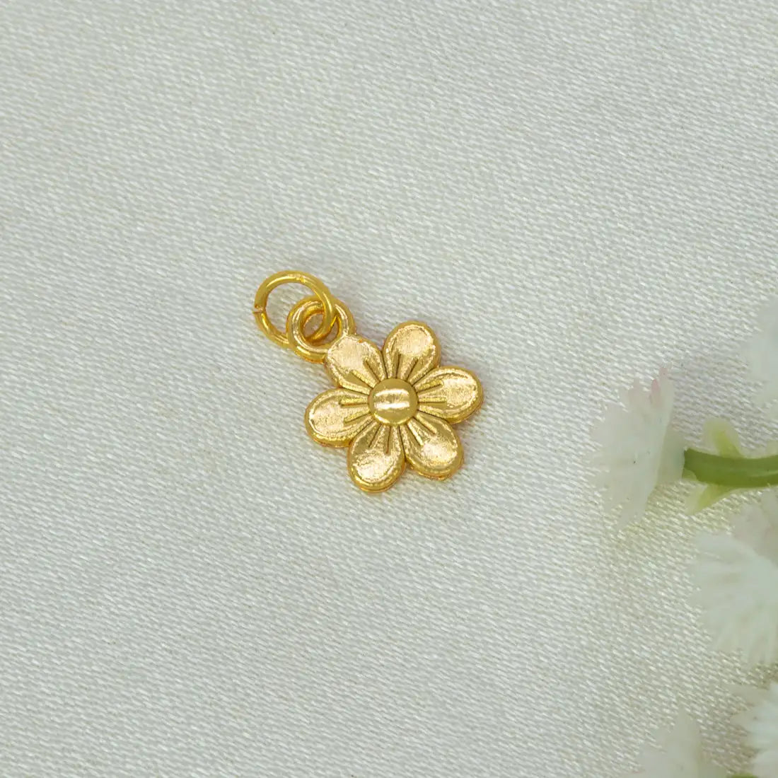 A Gold-Plated Textured Flower Pendant Charm with a textured center and a loop at the top, placed on a light textured fabric background with soft white flowers at the edge.