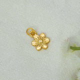 A Gold-Plated Textured Flower Pendant Charm with a textured center and a loop at the top, placed on a light textured fabric background with soft white flowers at the edge.