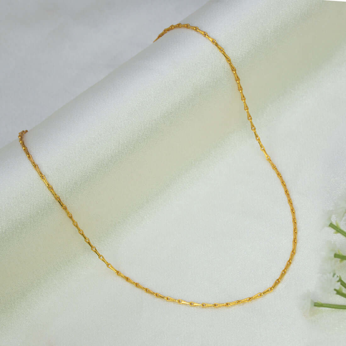 A delicate Gold-Plated Thin Baby Gopi Chain - 15 inches draped in a U-shape over a soft, off-white fabric background with a small white flower prop in the corner.