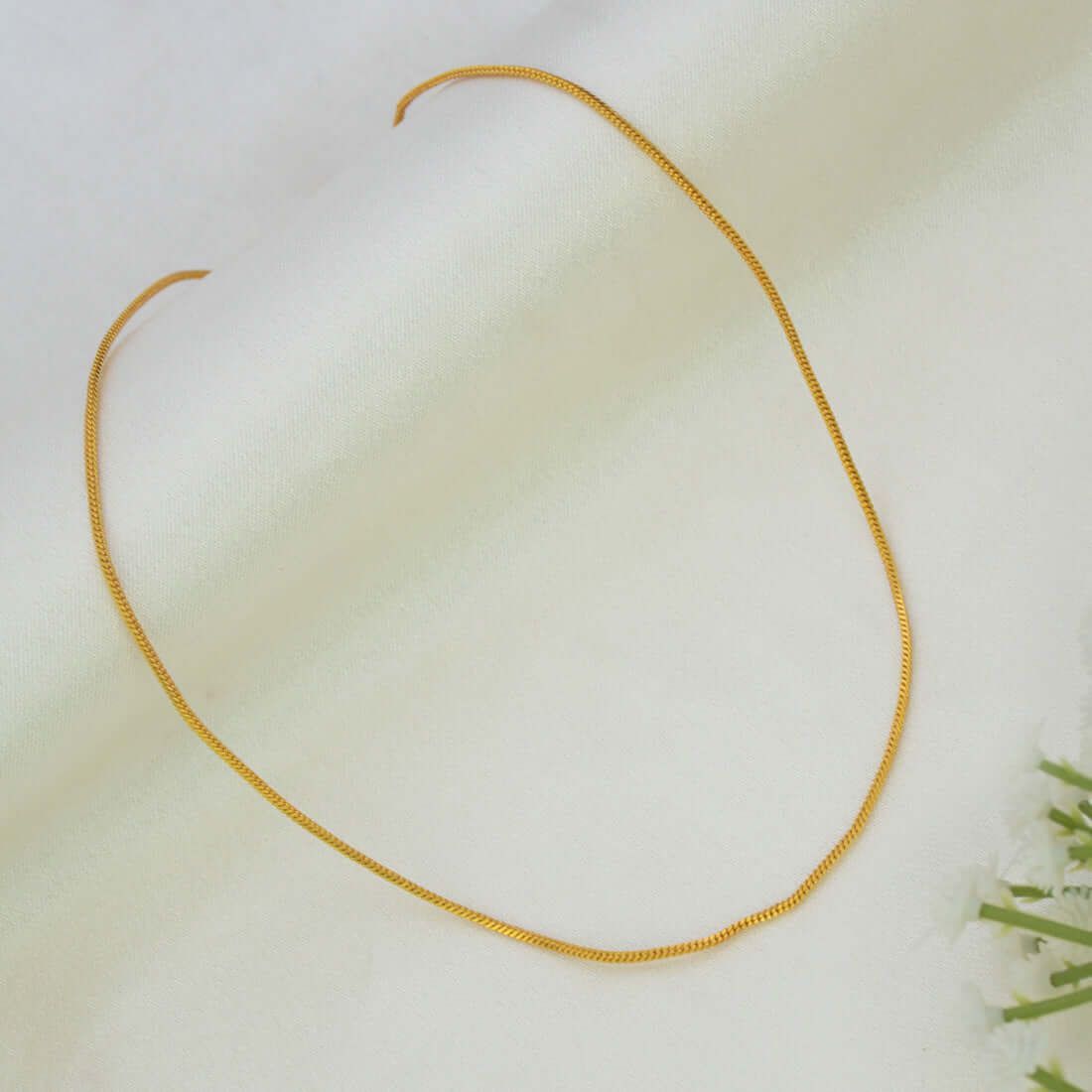 A delicate Gold-Plated Thin Snake Baby Chain necklace arranged in an oval shape on a soft, off-white textured fabric background with a hint of green leaves in the bottom right corner.