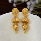 A pair of Gold-Plated Three-Step Filigree Jhumka Earrings displayed on a white fabric jewelry stand, with a soft-focus background of white flowers.