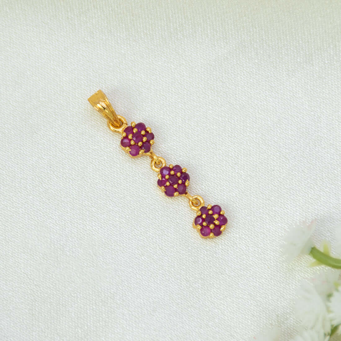 A Gold-Plated Three-tier Stone Floral Drop Pendant featuring three small ruby flower clusters connected in a vertical dangle design, displayed on a textured white fabric background.