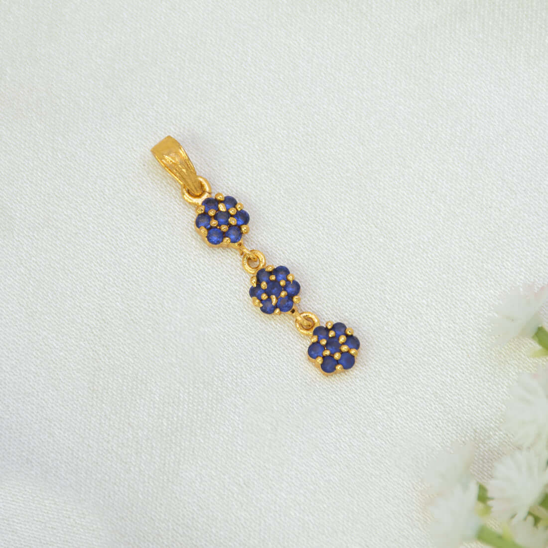A Gold-Plated Three-tier Stone Floral Drop Pendant featuring three small flower clusters with deep blue stones, displayed on a textured white fabric background with white flowers at the corner.