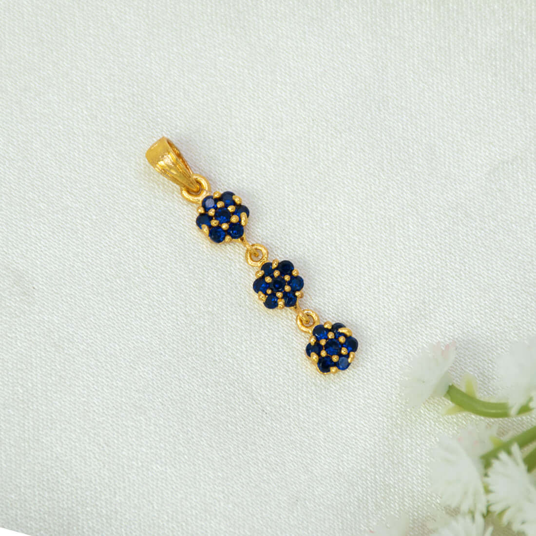 A Gold-Plated Three-tier Stone Floral Drop Pendant charm featuring three small flower clusters, each detailed with deep blue sapphire stones and gold accents, resting on a textured white fabric background with white flowers in the corner.