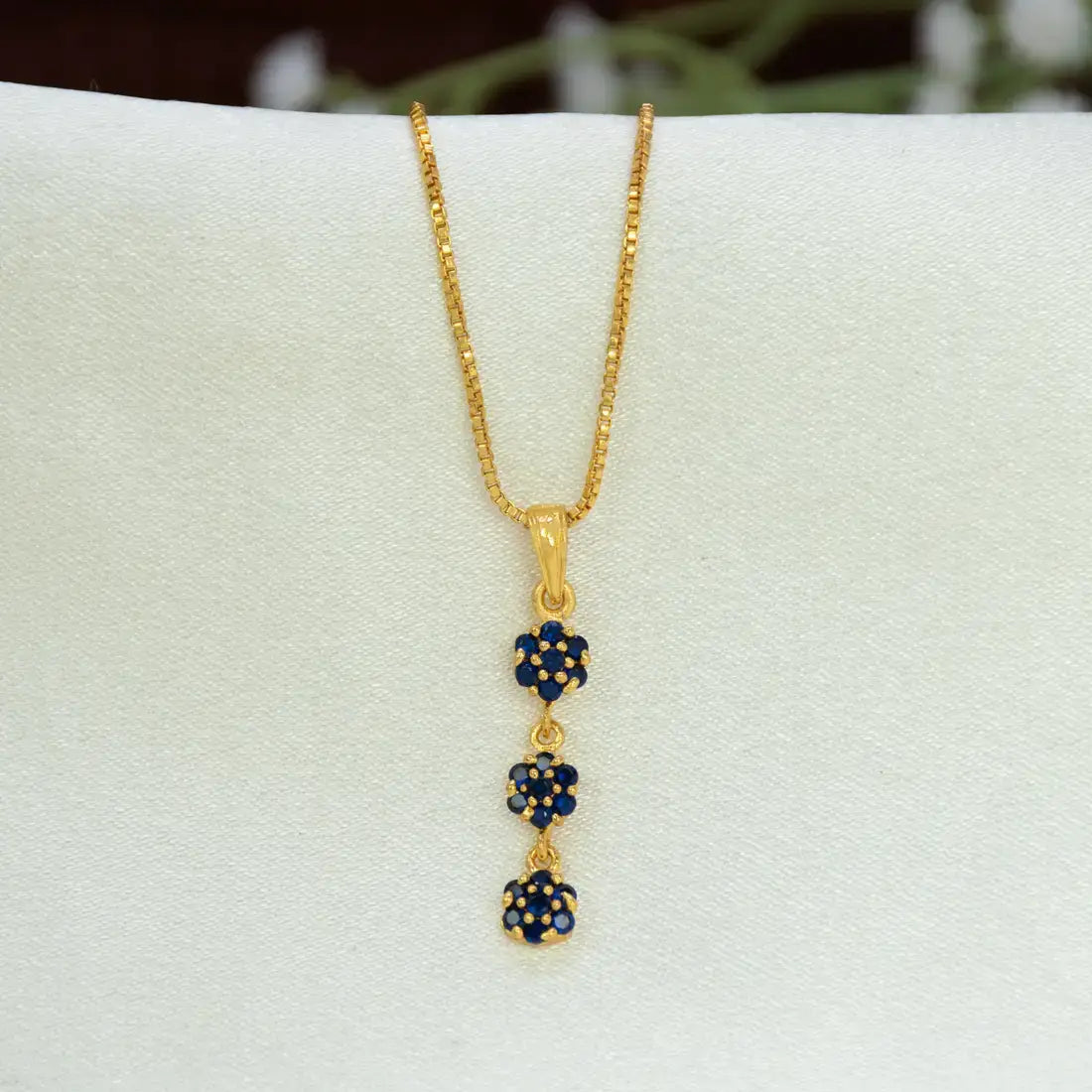 A gold chain necklace with a vertical Gold-Plated Three-tier Stone Floral Drop Pendant featuring three small flower-like clusters of deep blue sapphire stones set in gold, displayed on a white jewelry stand.