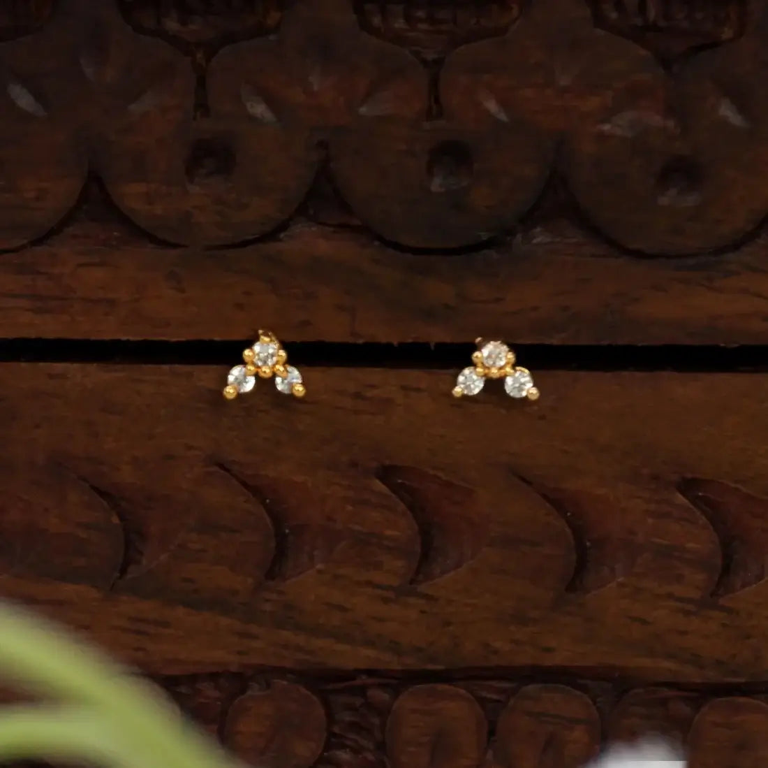 A pair of Gold-Plated Triple CZ Stone Second Stud Earrings with clear gemstones, displayed on a carved dark brown wooden surface.
