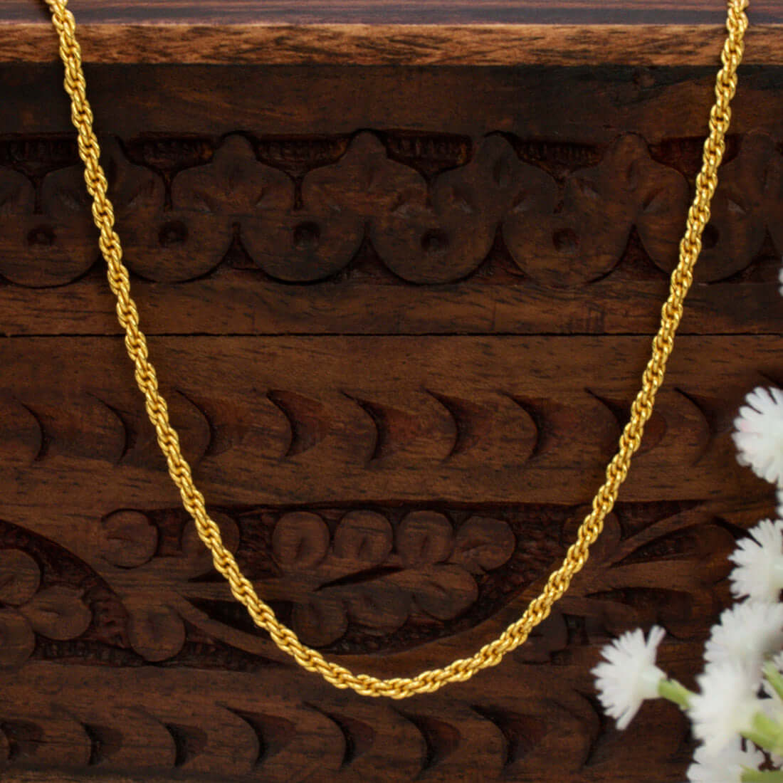 A Gold-Plated Twisted Anjali Baby Chainnecklace draped over a dark brown carved wooden box, with small white flowers in the bottom right corner.