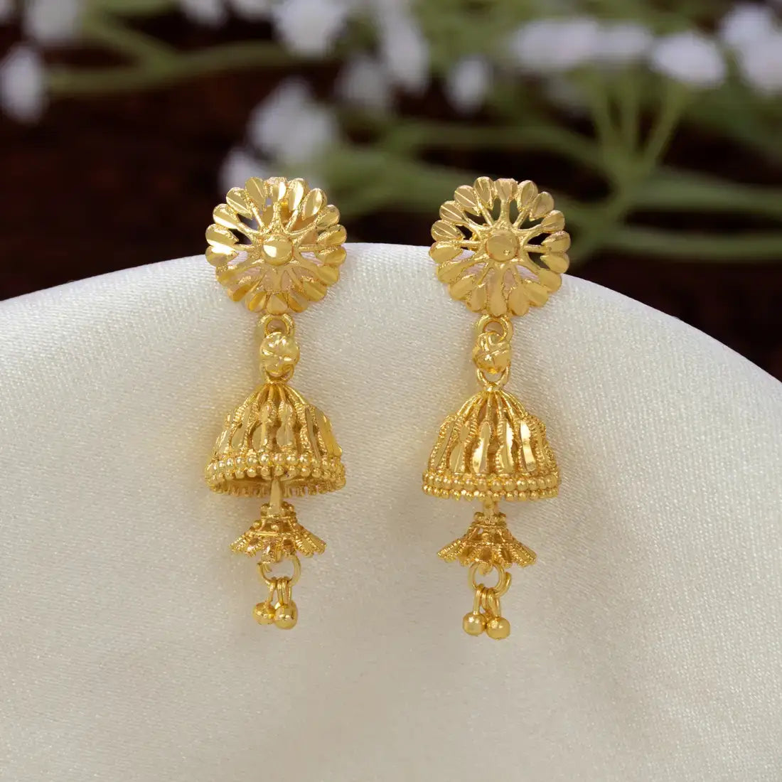 A pair of Gold-Plated Two-Step Floral Jhumka Earrings displayed on a white fabric stand. The earrings feature an intricate floral stud top and a dangling bell-shaped filigree dangle with small bead accents.
