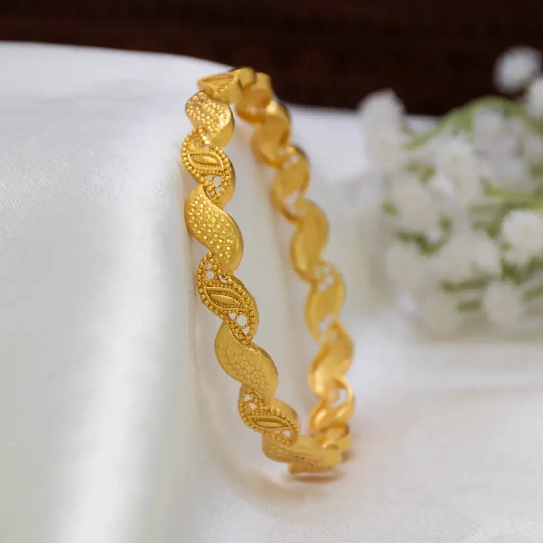 A Gold-Plated Wave Leaf Designer Bangle For Women with textured and patterned links, displayed on a draped white fabric surface with small white flowers in the background.