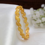 A Gold-Plated Wave Leaf Designer Bangle For Women with textured and patterned links, displayed on a draped white fabric surface with small white flowers in the background.