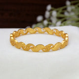 A Gold-Plated Wave Leaf Designer Bangle For Women with a textured, wavy interlocking link design, displayed on a white surface with a soft-focus background.