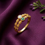 A gold ring with an intricate leaf-patterned band, featuring a clear round gemstone and a green oval gemstone, displayed on a draped purple fabric background.