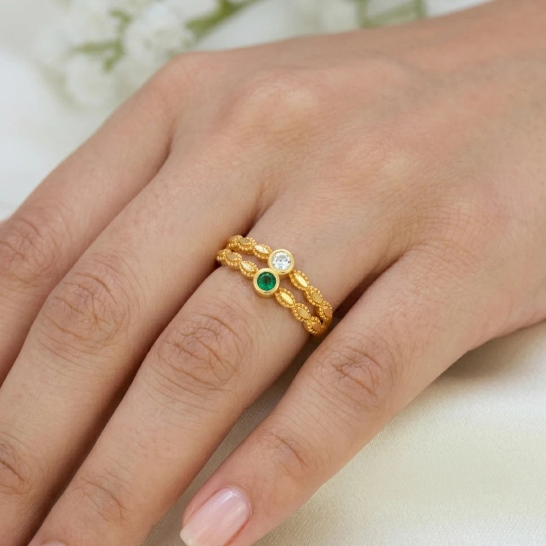 A close-up of a hand wearing a delicate gold ring with three small circular stones, including one green stone and one clear stone, set against a soft light background.