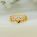 A gold-plated ring with a textured, braided filigree design, featuring a clear round-cut stone on one side and a small green oval stone on the other, resting on a soft white fabric background.