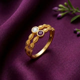 A gold ring with intricate leaf-like detailing, featuring a clear round gemstone and a round purple gemstone, displayed on a draped purple fabric background.