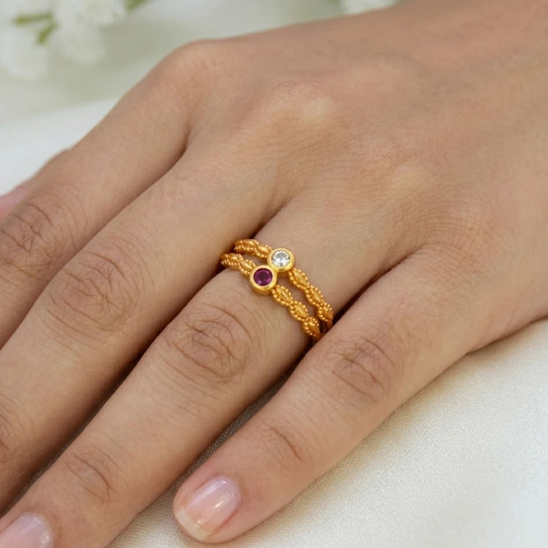 A close-up of a hand wearing a delicate gold ring with two twisted bands and small round gemstones, one clear and one purple, on a soft light background.