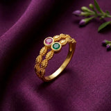 A gold ring with an intricate textured design and two round gemstones, one pink and one green, displayed on a draped purple fabric background.