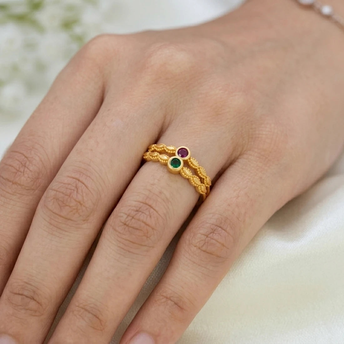 A close-up of a hand wearing a gold ring with two small round gemstones, one green and one red, on a soft light background.