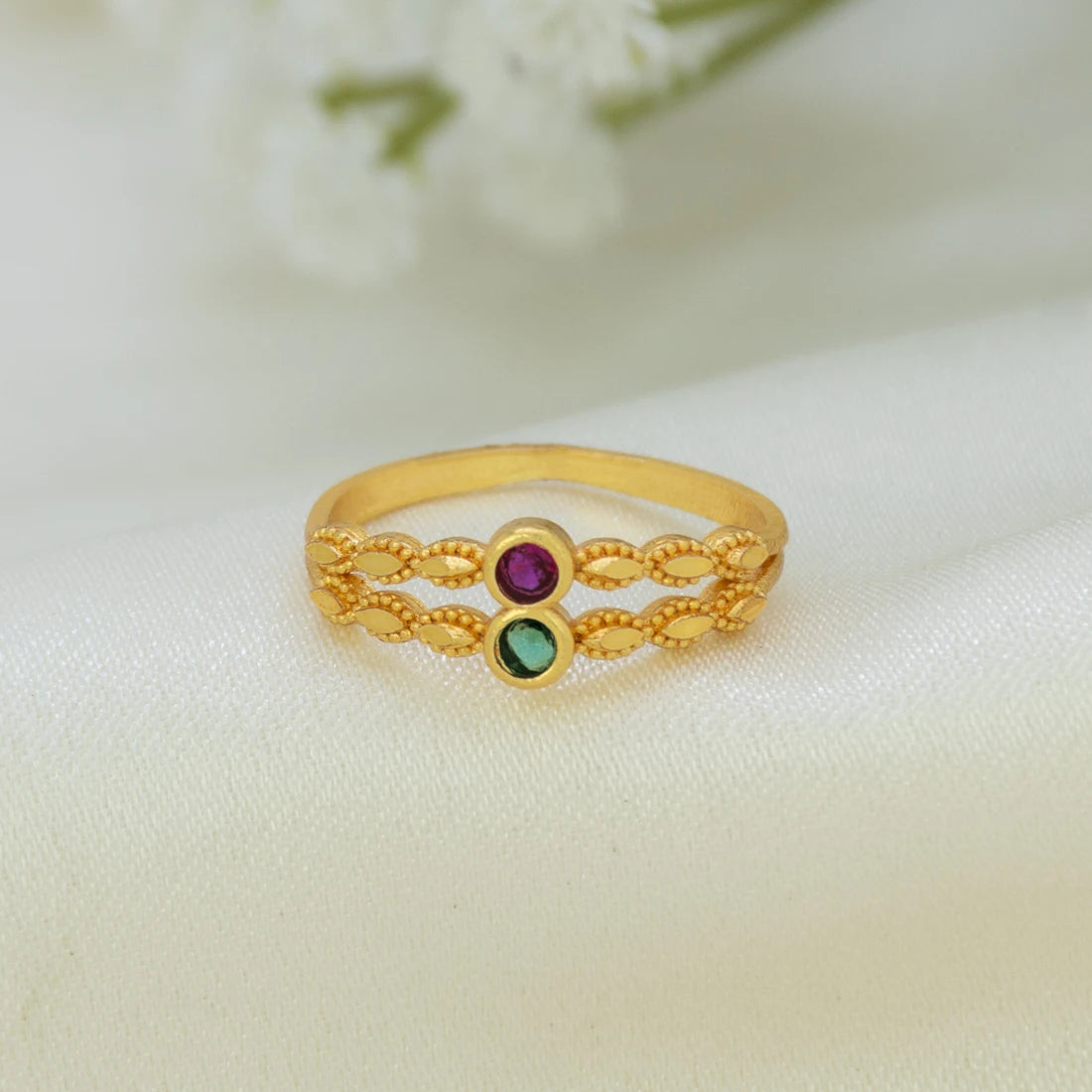 A gold ring with a textured chain-link design and two small round gemstones, one purple and one green, displayed on a soft white fabric background.