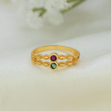 A gold ring with a textured chain-link design and two small round gemstones, one purple and one green, displayed on a soft white fabric background.