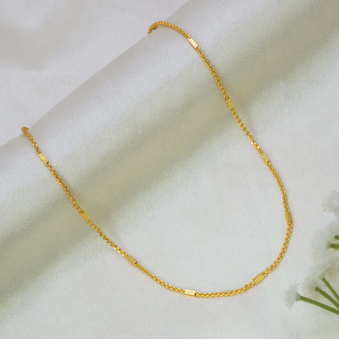 A delicate Gold Plated Designer Box Cutting Baby Chain with small flat rectangular links, displayed on a soft white fabric background with a small white flower in the corner.